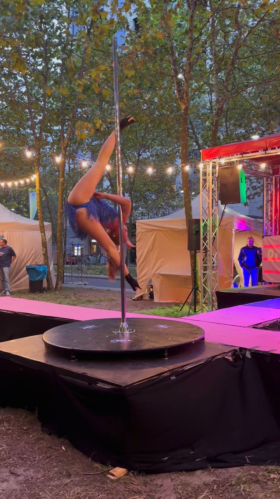 Megan Konings performing an inverted split on a stage pole at an outdoor festival at dusk