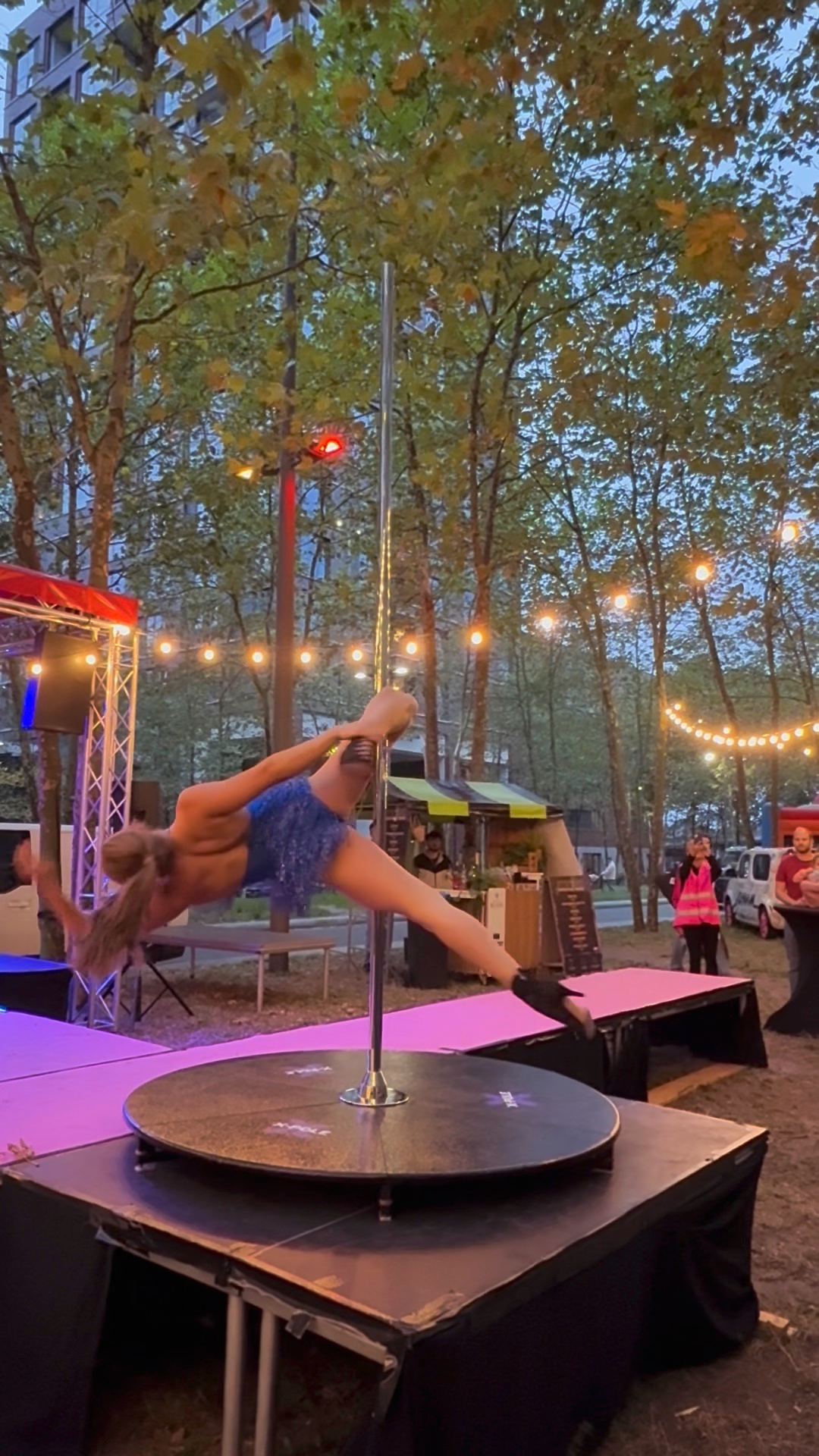 Megan spinning on the pole in a sparkling blue costume, festival lights behind her